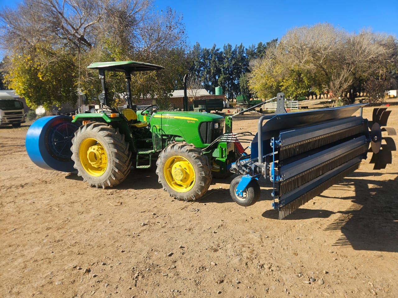 Tractor fitted with a rear-mounted blue blower and a front-mounted sweeping attachment in a farm yard