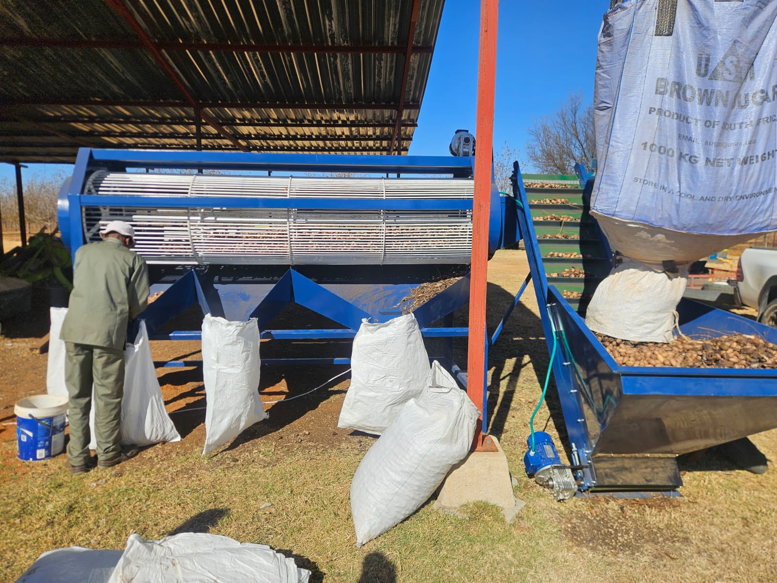 Blue and white nut-processing machine with a conveyor and bagging chutes running under a metal roof
