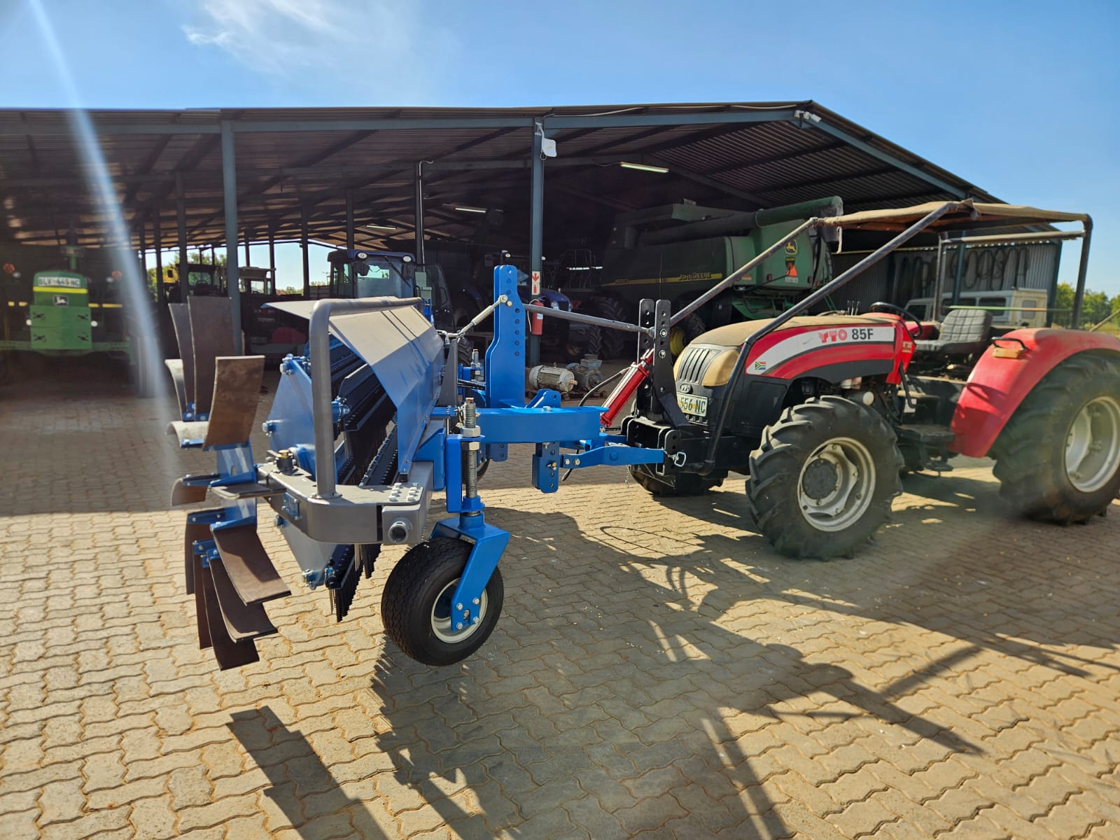 Red tractor with a blue front-mounted rake and sweeper beside an open machinery shed